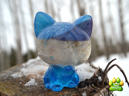 Petit chat bleu avec paillette et pissenlit integres sur un tronc d'arbre enneigé avec foret flouter arriere 